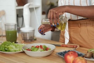 Mani di giovane femmina che versa olio d'oliva nell'insalata di verdure mentre si trova vicino al tavolo della cucina