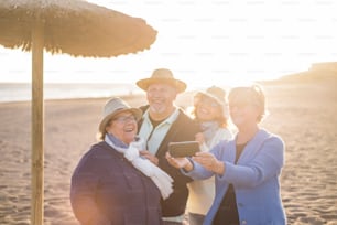 elderly senior group of friends with a 40s woman having phone at the beach with a mobile phone. Sunlight in backlight while they take a selfie.