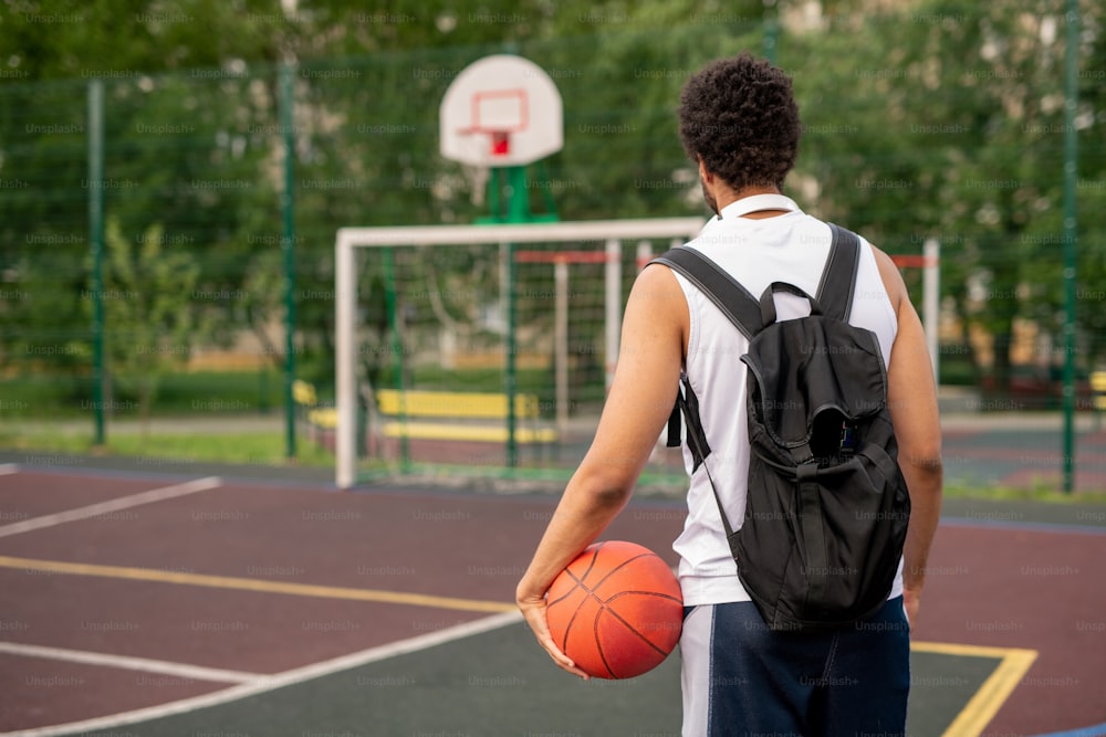 Rear view of young sportsman with black backpack and ball coming to basketball court to play