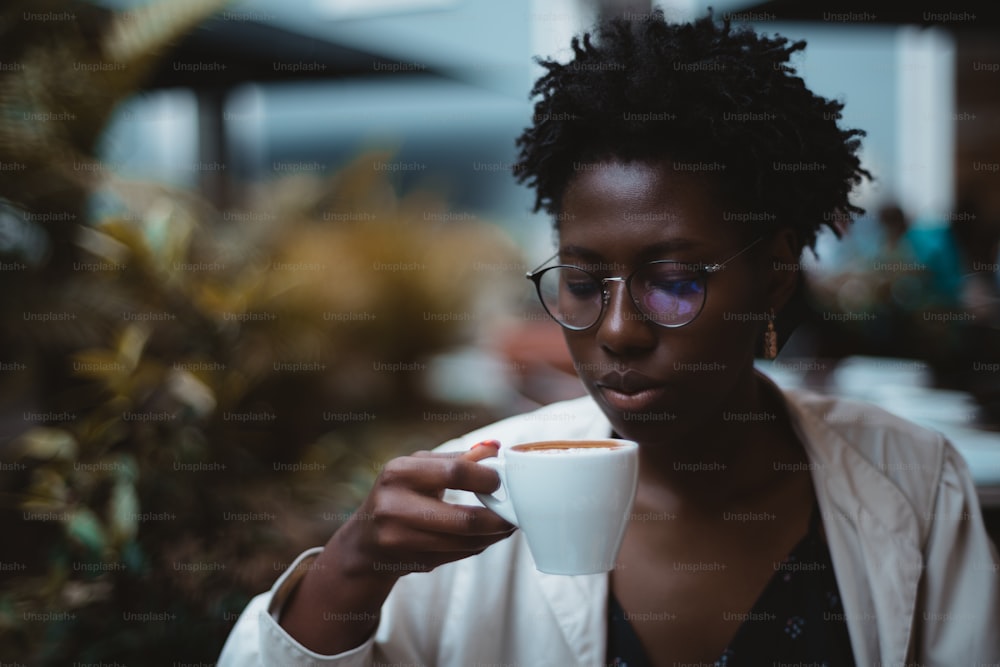 A low-key portrait of a charming young black female with afro curls, in elegant glasses and a white trench coat, drinking a delicious hot chocolate in a dark night cafe; a copy space place on the left