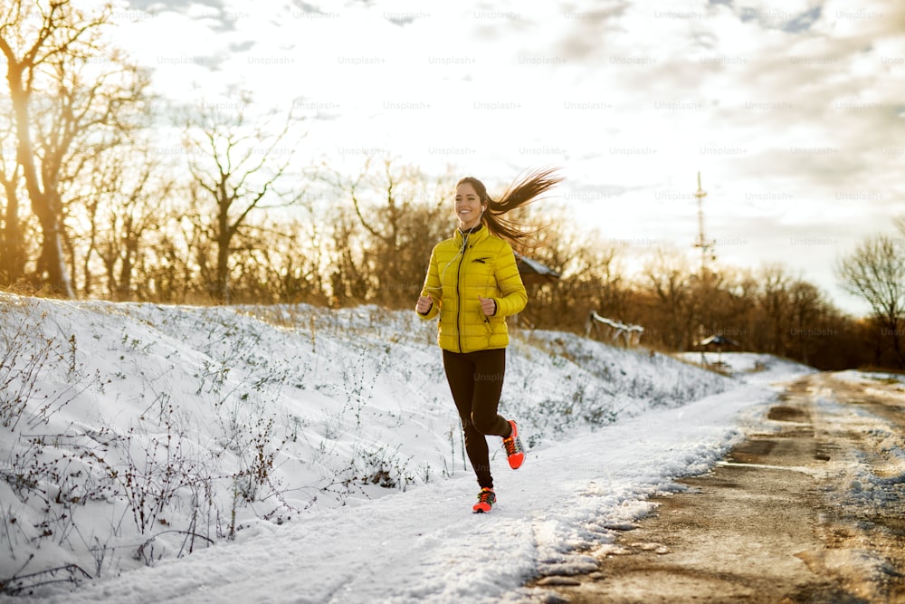 Young attractive healthy runner girl jogging in winter sportswear on snowy winter road outside with earphones in the sunny morning.