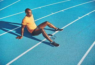 Fit young African runner sitting alone with his cellphone on a track preparing for a run on a sunny day