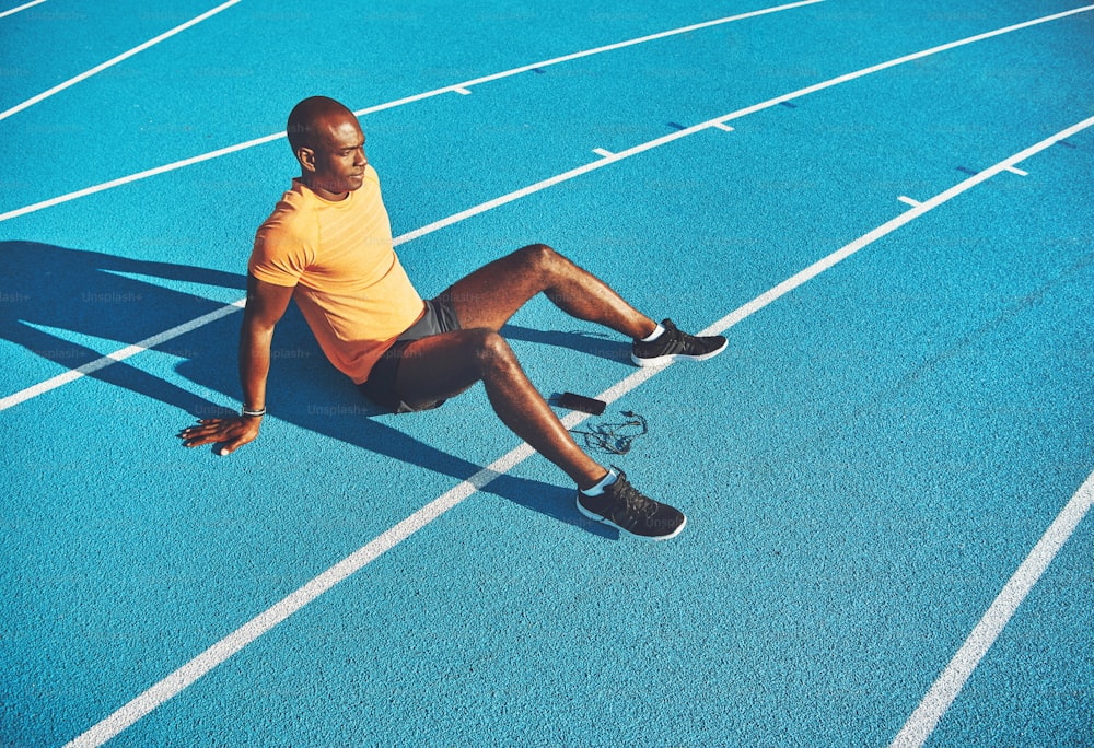 Fitter junger afrikanischer Läufer, der allein mit seinem Handy auf einer Strecke sitzt und sich auf einen Lauf an einem sonnigen Tag vorbereitet