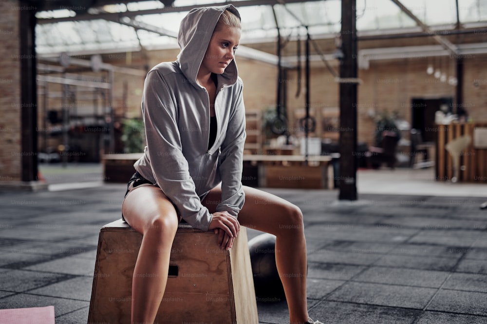 Sportliche junge blonde Frau im Kapuzensweatshirt sitzt nach dem Training im Fitnessstudio auf einer Kiste