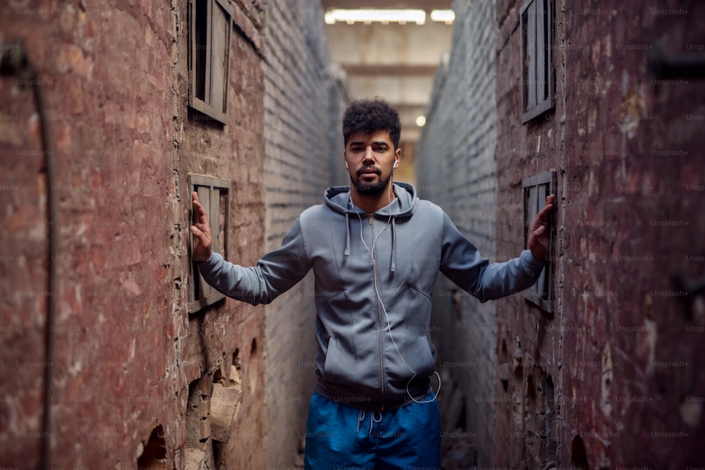 Portrait of focused motivated afro-american young handsome sportive man with earphones standing inside of the abandoned place in the middle of two walls and thinking while looking at camera.