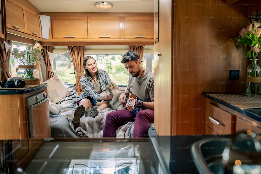 Young man playing guitar while his girlfriend cuddling husky dog inside travel house