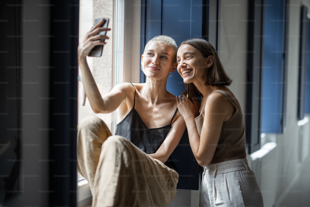 Cheerful girlfriends or lesbian couple have fun, making selfie photo or talking online by cell phone sitting by the window indoors