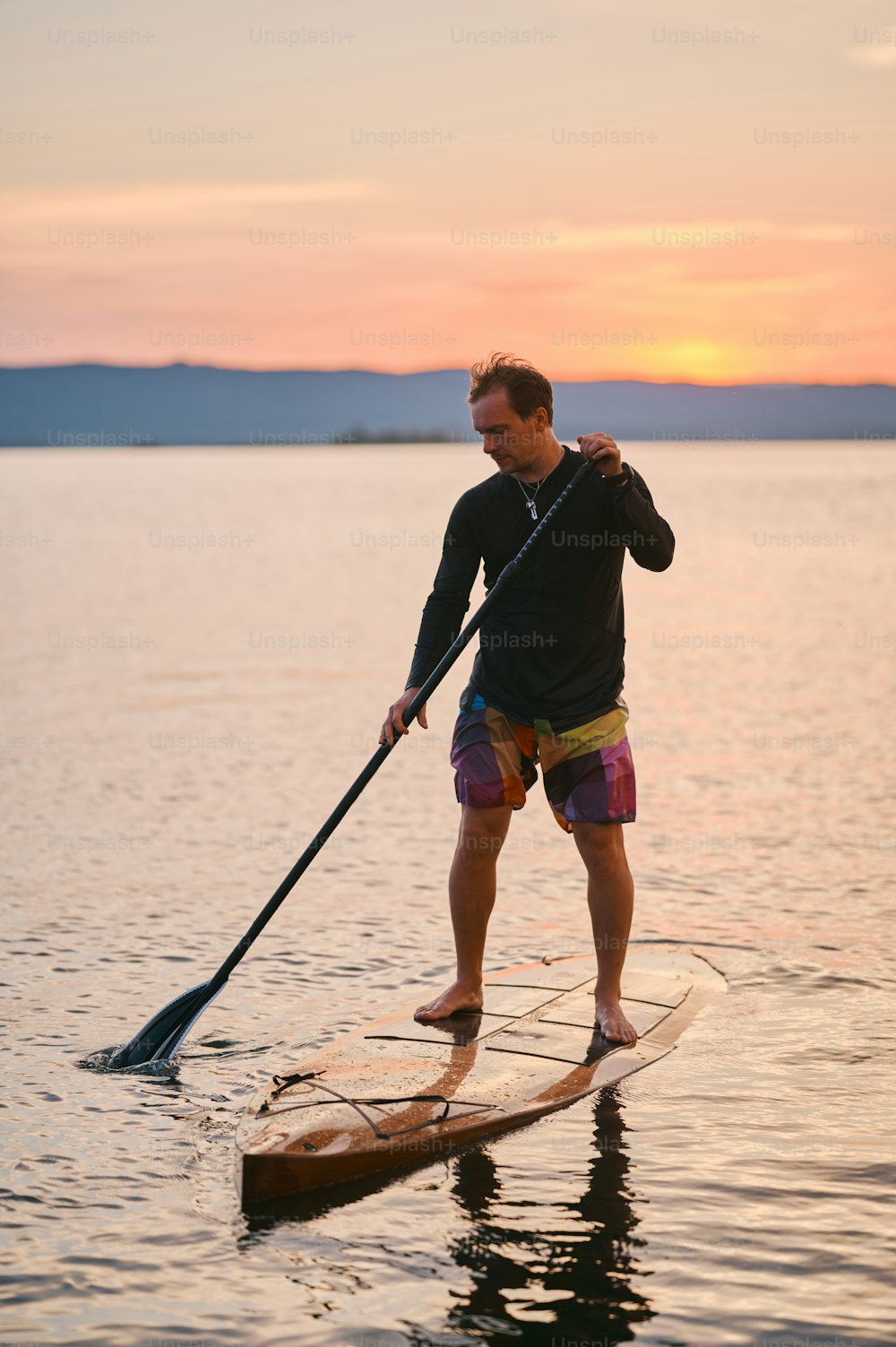 Ganzkörperaufnahme eines Mannes mit Paddel, der auf Holzbrett in ruhigem Wasser steht und bei Sonnenuntergang mit malerischem Bergblick surft