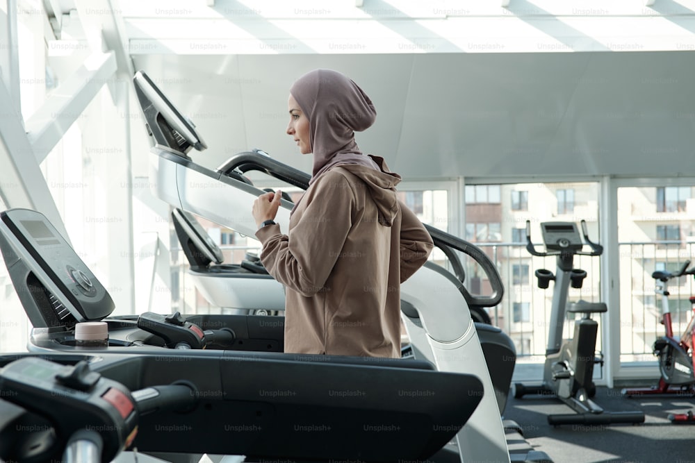 히잡과 운동복을 입은 젊은 활동적인 여성이 체육관에서 여가를 보내는 동안 러닝머신에서 뛰고 있다