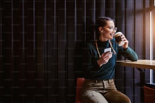 Smiling teenage girl sitting in cafeteria next to window, drinking coffee and holding smart phone while looking trough window.