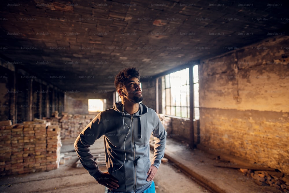 Retrato de un joven afroamericano guapo y deportivo con auriculares de pie dentro del lugar abandonado. Retrato exitoso de un joven activo.
