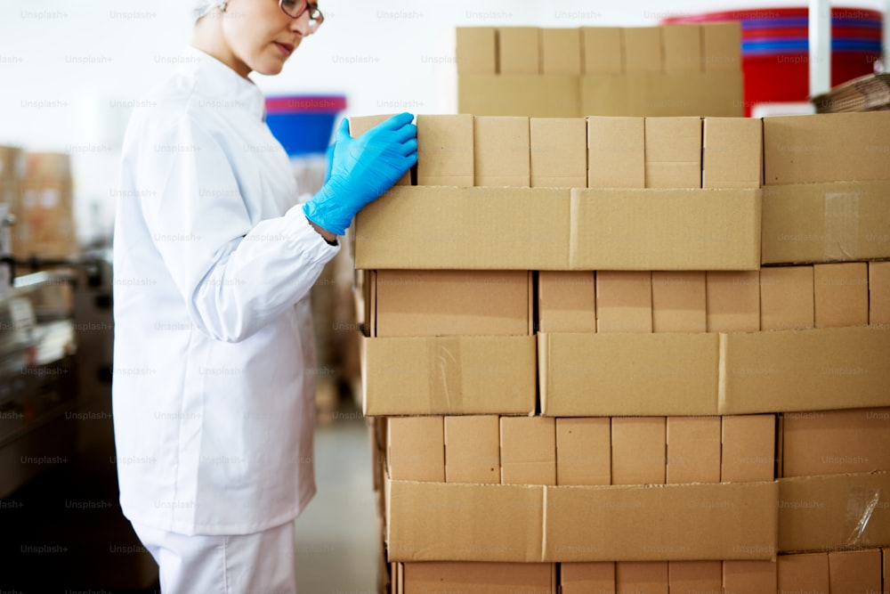 Young focused female worker in sterile cloths is folding boxes on stacks of boxes in factory storage room.