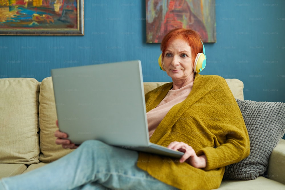Elderly woman in wireless headset sitting on sofa with laptop on her knees and watching movie online during leisure time at home