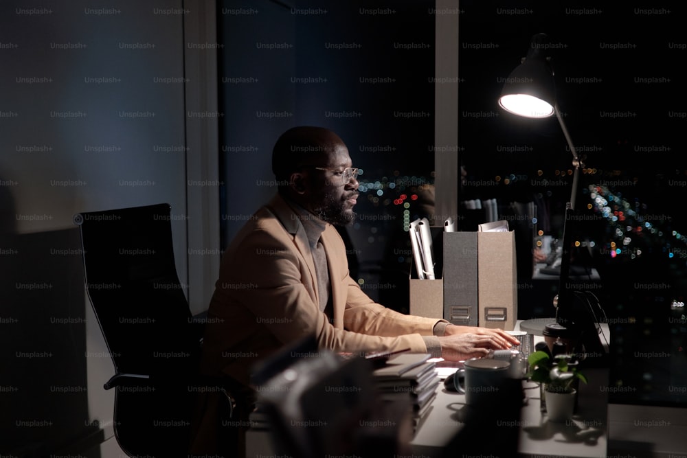 바쁜 젊은 아프리카 남자가 컴퓨터 키보드에 타이핑하고 사무실에서 늦게까지 일하는 동안 화면을 보고 있다