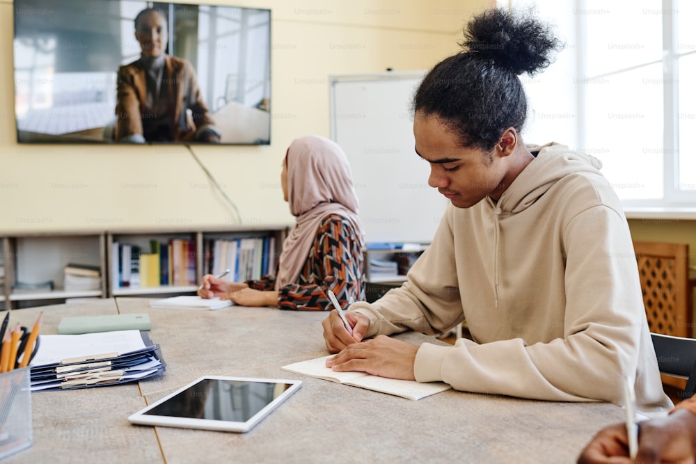 Des élèves multiethniques assis à table regardent des vidéos éducatives et prennent des notes dans des cahiers pendant la leçon