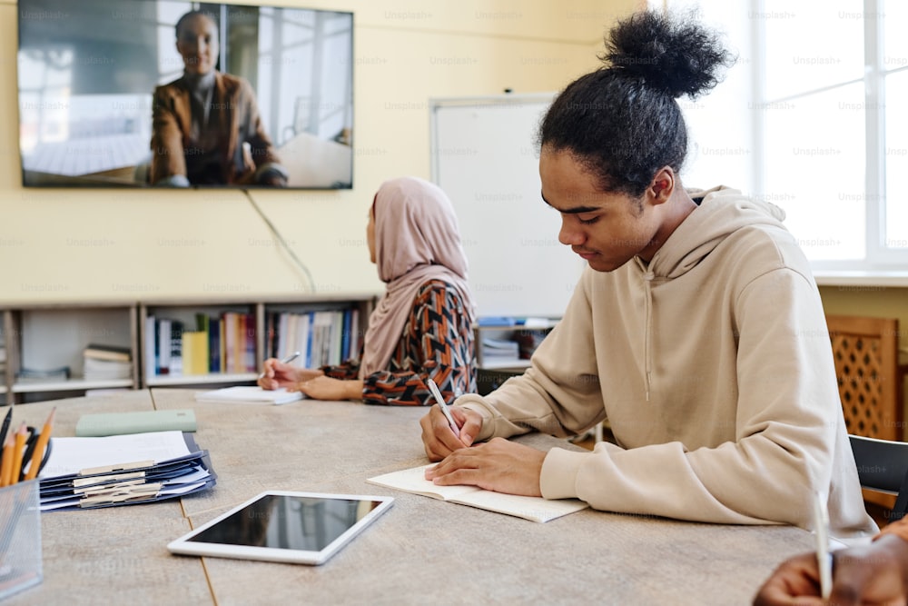 다민족 학생들이 테이블에 앉아 교육 비디오를 보고 수업 중 노트북에 메모를 합니다.