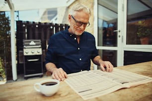 Smiling senior man sitting at his patio table outside reading the financial pages in a newspaper and drinking a cup of coffee