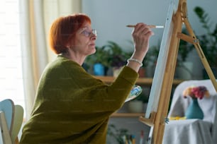Elderly woman in eyeglasses concentrating on her painting on easel during her leisure time at home