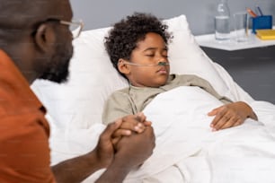訪問中に父親が手を握って病棟のベッドで寝ているアフリカの小さな男の子
