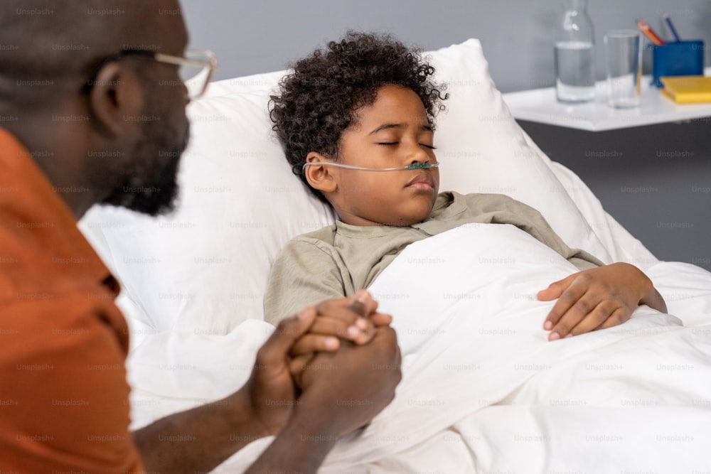 Afrikanischer kleiner Junge schläft im Bett auf der Krankenstation mit Vater, der während des Besuchs seine Hand hält