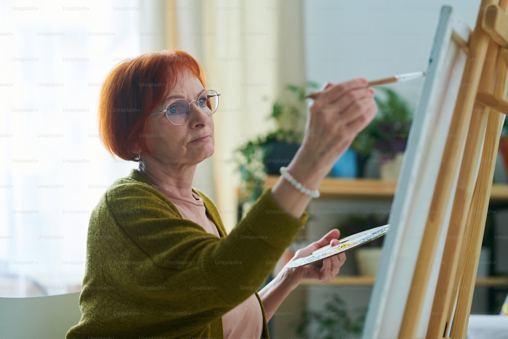 Mujer mayor activa sosteniendo paleta y pincel y pintando sobre lienzo en la habitación de su casa