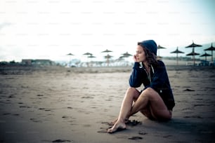 Süßes, einsames kaukasisches junges Mädchen, das am Strand im Sand sitzt und den offenen Raum betrachtet und nachdenkt - geschlossenes Resort mit Sonnenschirmen im Hintergrund - kalte Töne und Farben