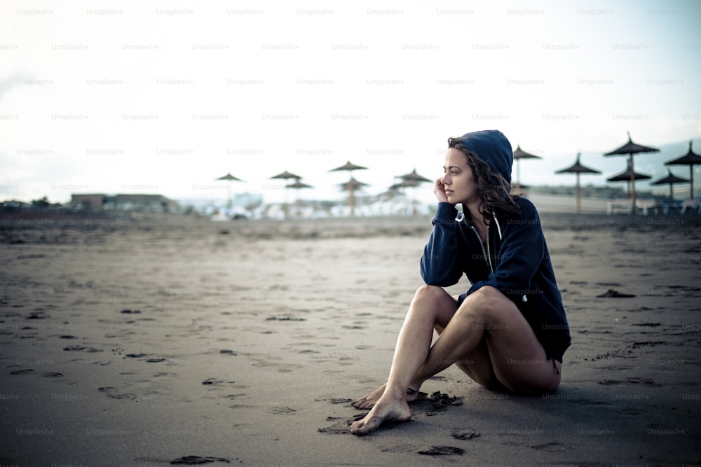Cute lonely caucasian young girl sitting at the beach on the sand looking the open space and thinking - closed resort with sun umbrellas in background - cold tones and colors
