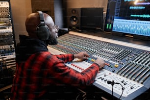 Stylish African American man wearing red checked shirt and headphones working on music track in recording studio