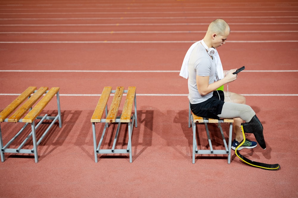 Jeune sportif avec prothèse sur la jambe droite assis sur le banc à la pause et à la recherche de quelque chose à écouter dans la playlist