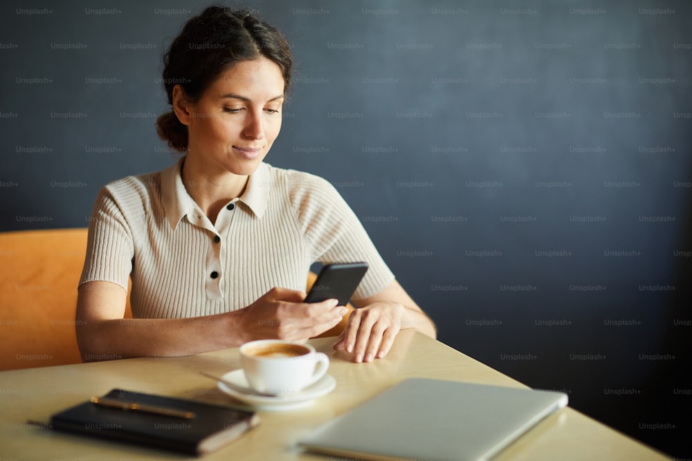 Joven empresaria seria desplazándose en un teléfono inteligente o enviando mensajes de texto a uno de los clientes mientras espera a alguien junto a la mesa en el café