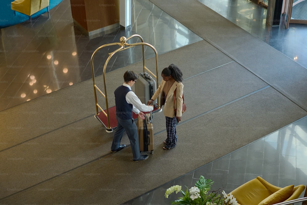 Above angle of young female traveler passing her suitcase to bellboy with cart standing in front of her in spacious lounge of hotel