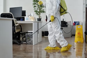 Jovem trabalhador do sexo masculino do serviço de limpeza em roupas de trabalho de proteção pulverizando piso entre locais de trabalho em escritório contemporâneo de espaço aberto
