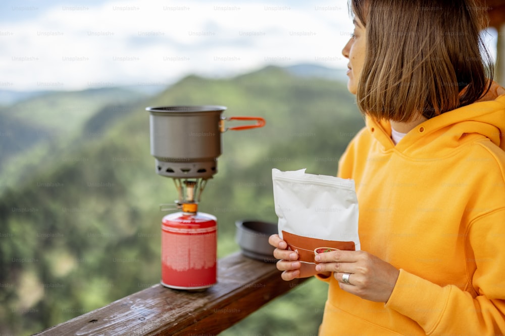 젊은 여성은 여름에 산을 여행하는 동안 가스 버너가 있는 밀폐 패키지로 하이킹을 위해 동결 건조 음식을 준비합니다. 하이킹 중 음식 개념