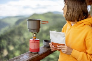 Young woman prepares freeze-dried food for hiking in airtight package, with gas burner, while traveling in mountains on summertime. Food concept during hiking
