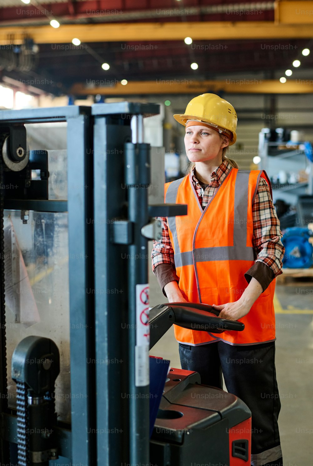 Female engineer looking forwards while standing in front of electric forklift and moving along aisle of workshop or warehouse