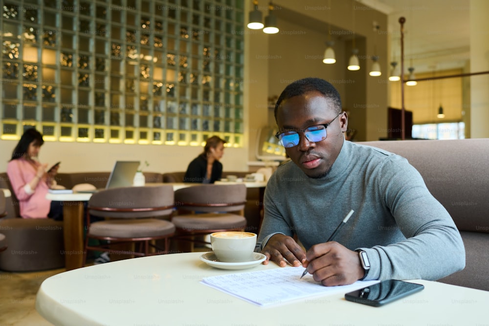 Joven hombre de negocios africano sentado a la mesa con café y tomando notas en el documento, él trabajando durante la pausa para el café en el café