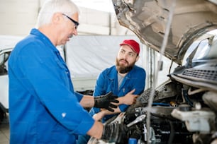 Young bearded worker of technical service looking at his mature colleague examining broken engine of large machine while listening to him