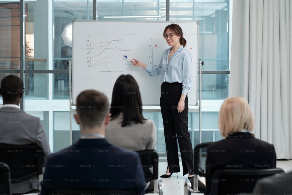 Happy young contemporary female economist pointing at graph on whiteboard during presentation of financial fluctuations