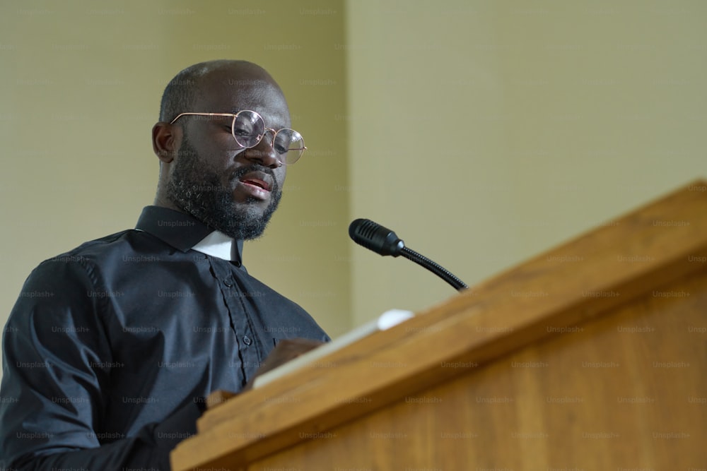 Young priest in black shirt with clerical collar reading verses from Bible and explaining them while pronouncing speech by pulpit