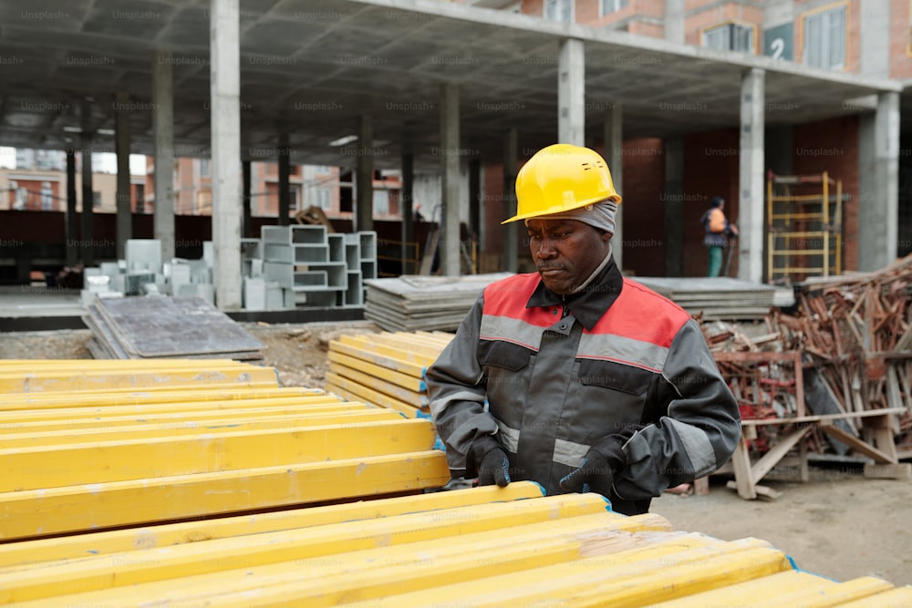 작업복을 입은 성숙한 흑인 남자가 노란 색의 건축 자재 더미 옆에 서서 건설 현장에서 일하는 동안 널빤지를 선택합니다.