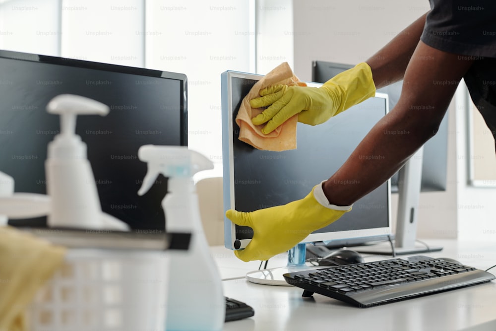Gloved hands of young African American man with duster wiping computer screen while standing by workplace of office manager
