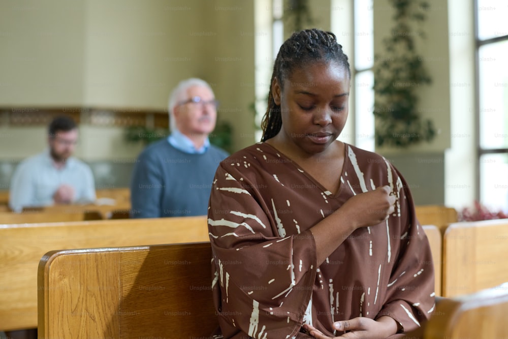 Young black woman with her eyes closed crossing herself during silent prayer while sitting on bench against parishioners in church