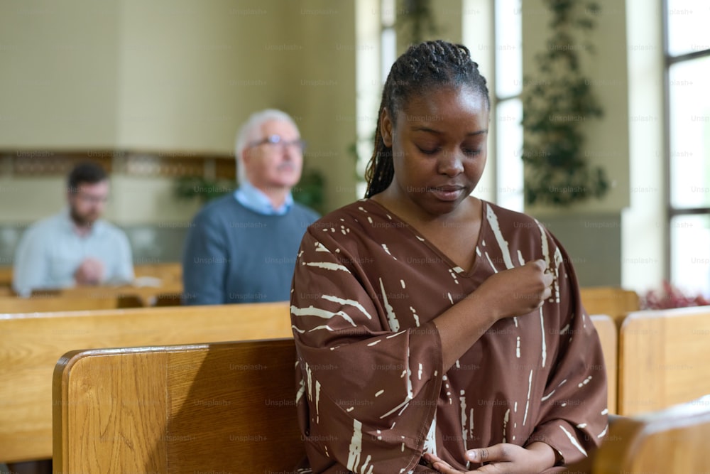 Junge schwarze Frau mit geschlossenen Augen bekreuzigt sich während des stillen Gebets, während sie auf der Bank gegen Gemeindemitglieder in der Kirche sitzt