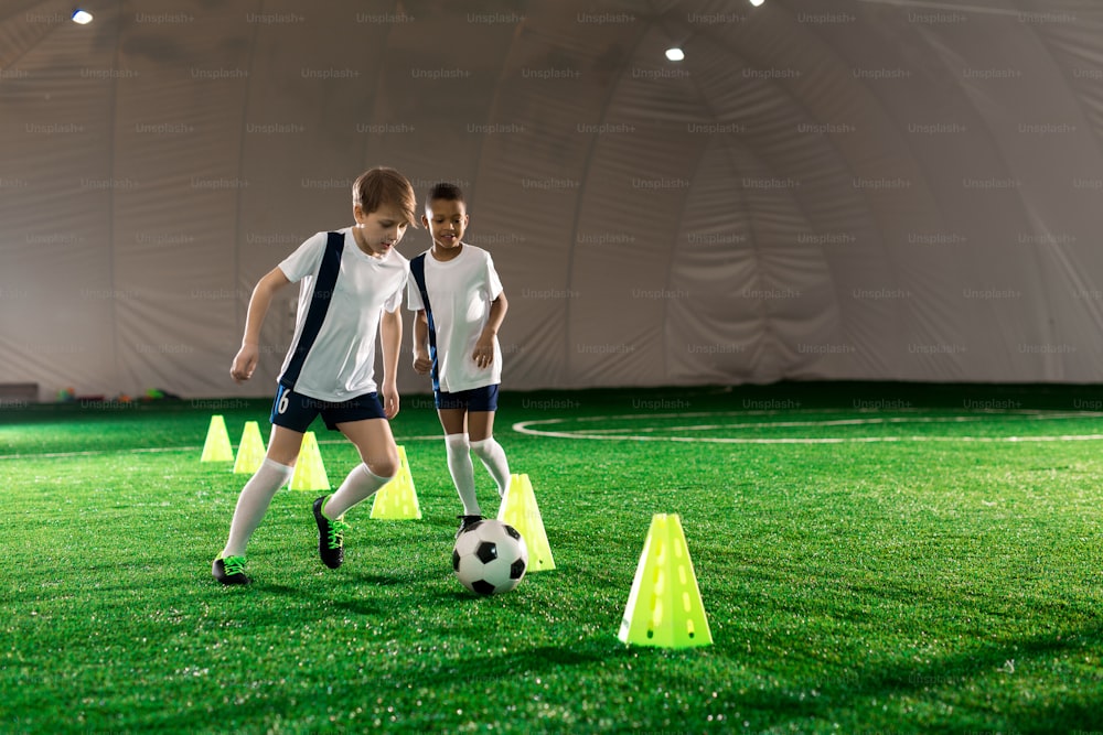 Two little football players running after ball while exercising on pitch with cones