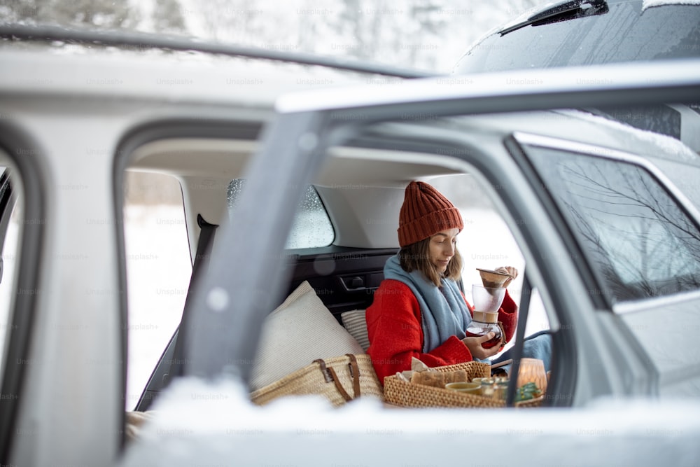 Frau trinkt Kaffee im Kofferraum und reist in den Winterferien mit dem Mietwagen. Blick aus dem Fenster, von außen ins Auto. Hochwertiges Foto