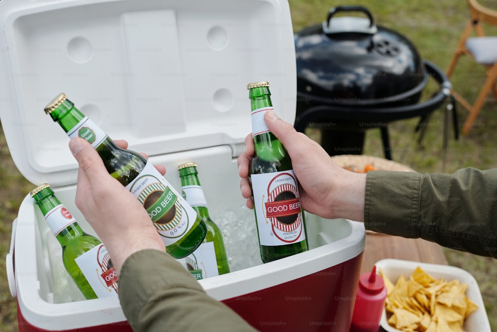 Hands of young man holding two bottles of beer over box prepared for guests and backyard party against barbecue grill and potato chips in bowl