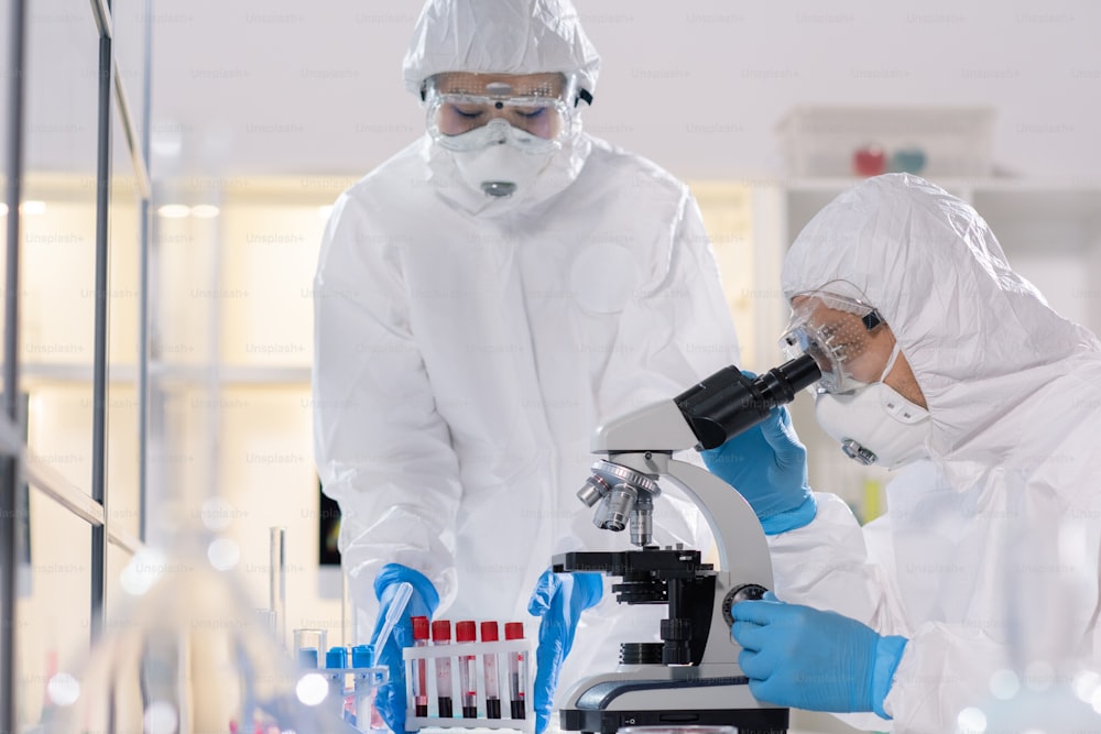 Joven científico en ropa de trabajo protectora mirando en el microscopio mientras estudia un nuevo virus con un colega en un laboratorio médico