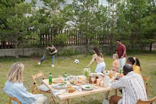 Jovem casal negro e mulher caucasiana sentada ao lado da mesa servida e olhando para dois homens e crianças interculturais jogando jogos ao ar livre