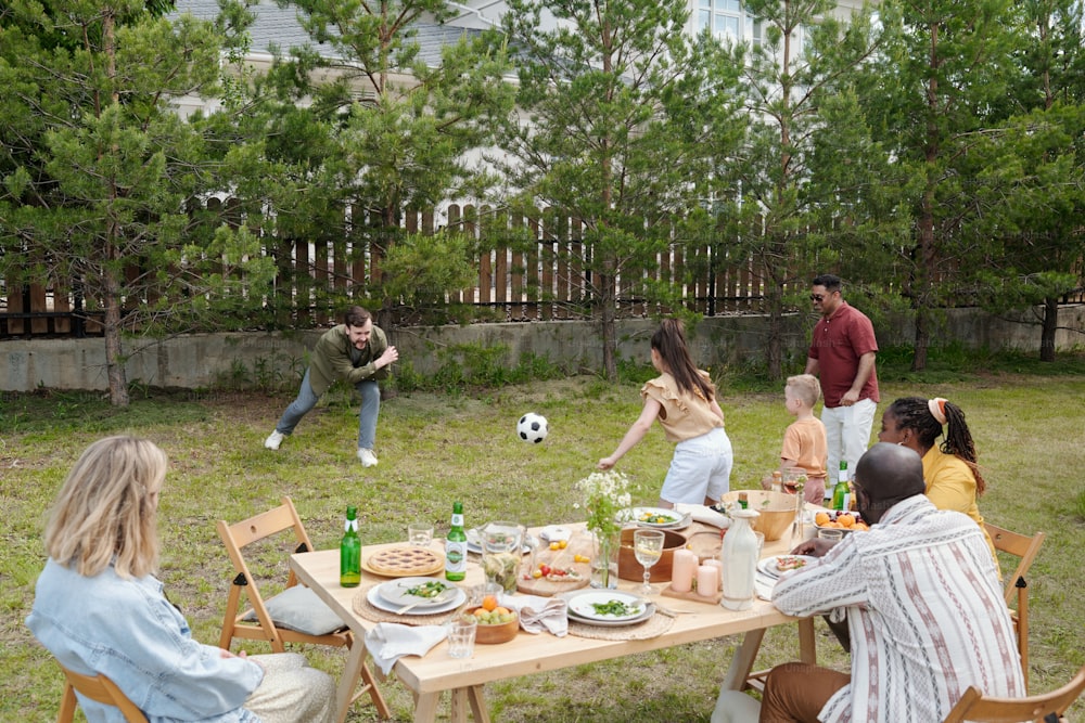 Young black couple and Caucasian woman sitting by served table and looking at two intercultural men and kids playing outdoor game