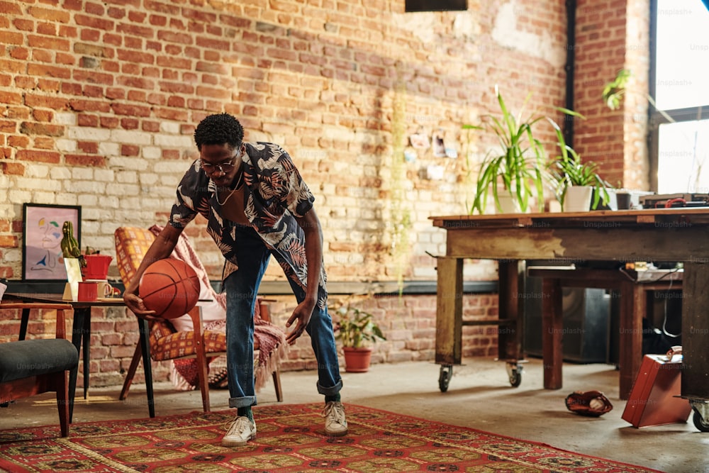 Young African American man in casualwear playing with ball on carpet in spacious living room of loft apartment with brick walls